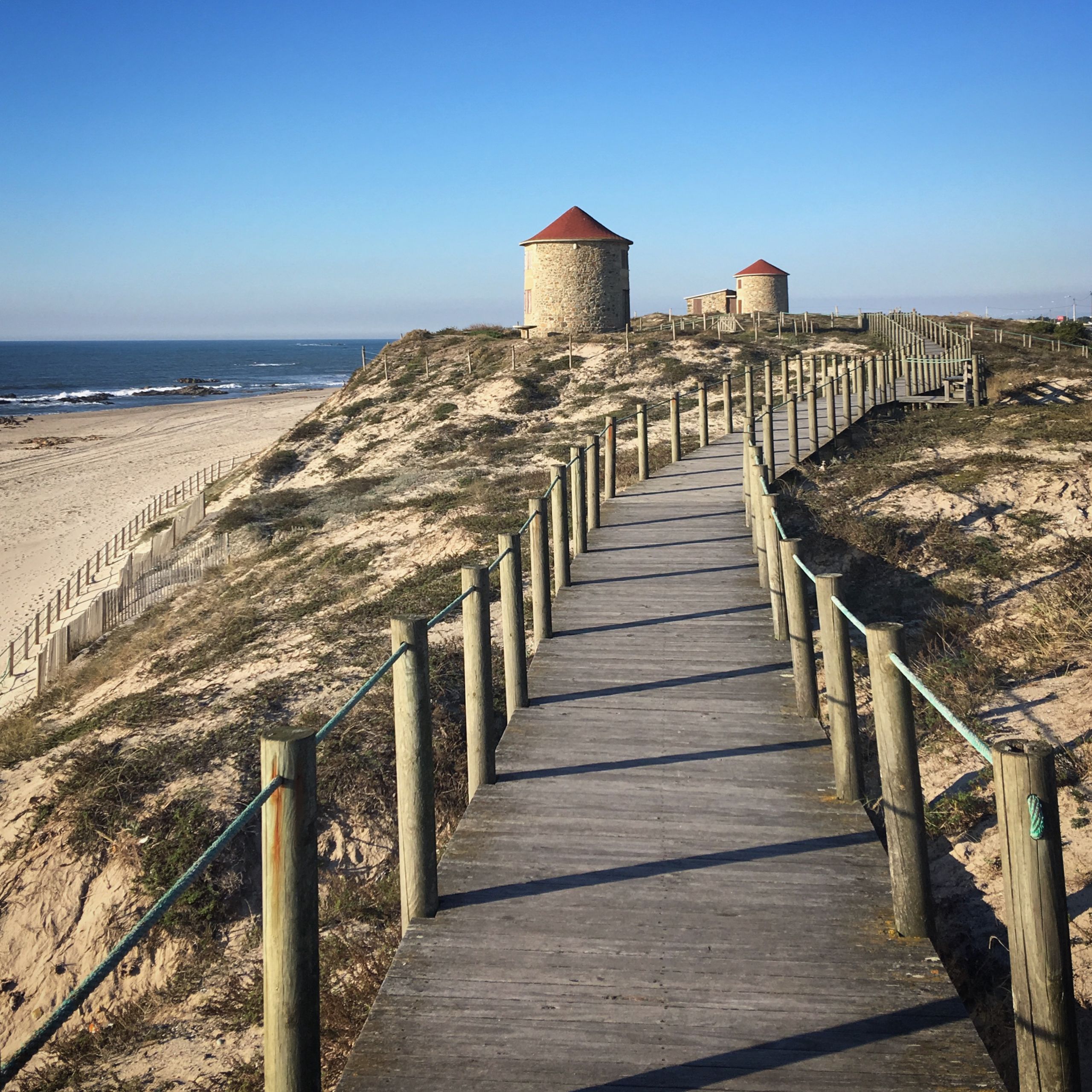A wooden boardwalk with scenic views of Apúlia beach and historic windmills under a clear blue sky.